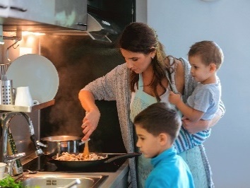 A mother cooking with one hand while holding her son with the other. Her other son is standing next to her.