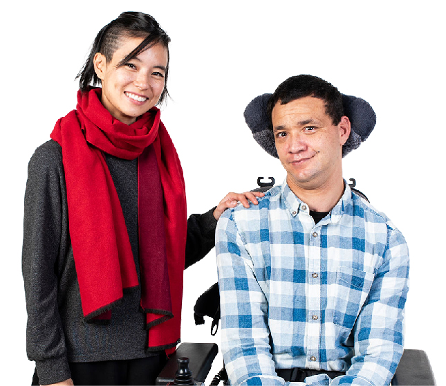 A person standing next to a person in a wheelchair. Both of them are smiling.